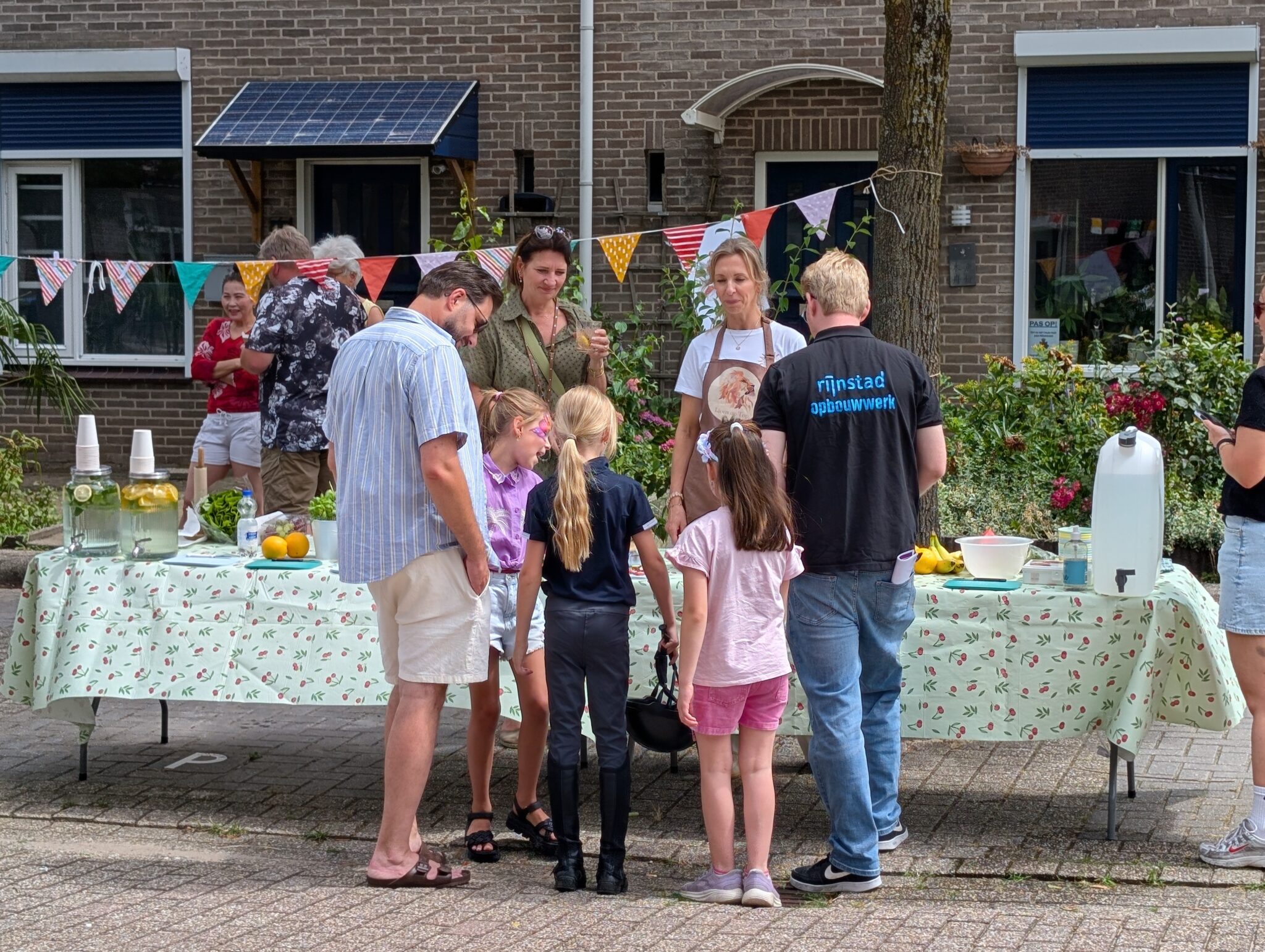 Buurfeest met limonadekraam, kleurrijke vlaggetjes en mensen die elkaar ontmoeten en praten.