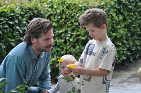 Vader leert kinderen over bloemen in een tuin. Een jongen houdt een gele bloem vast.