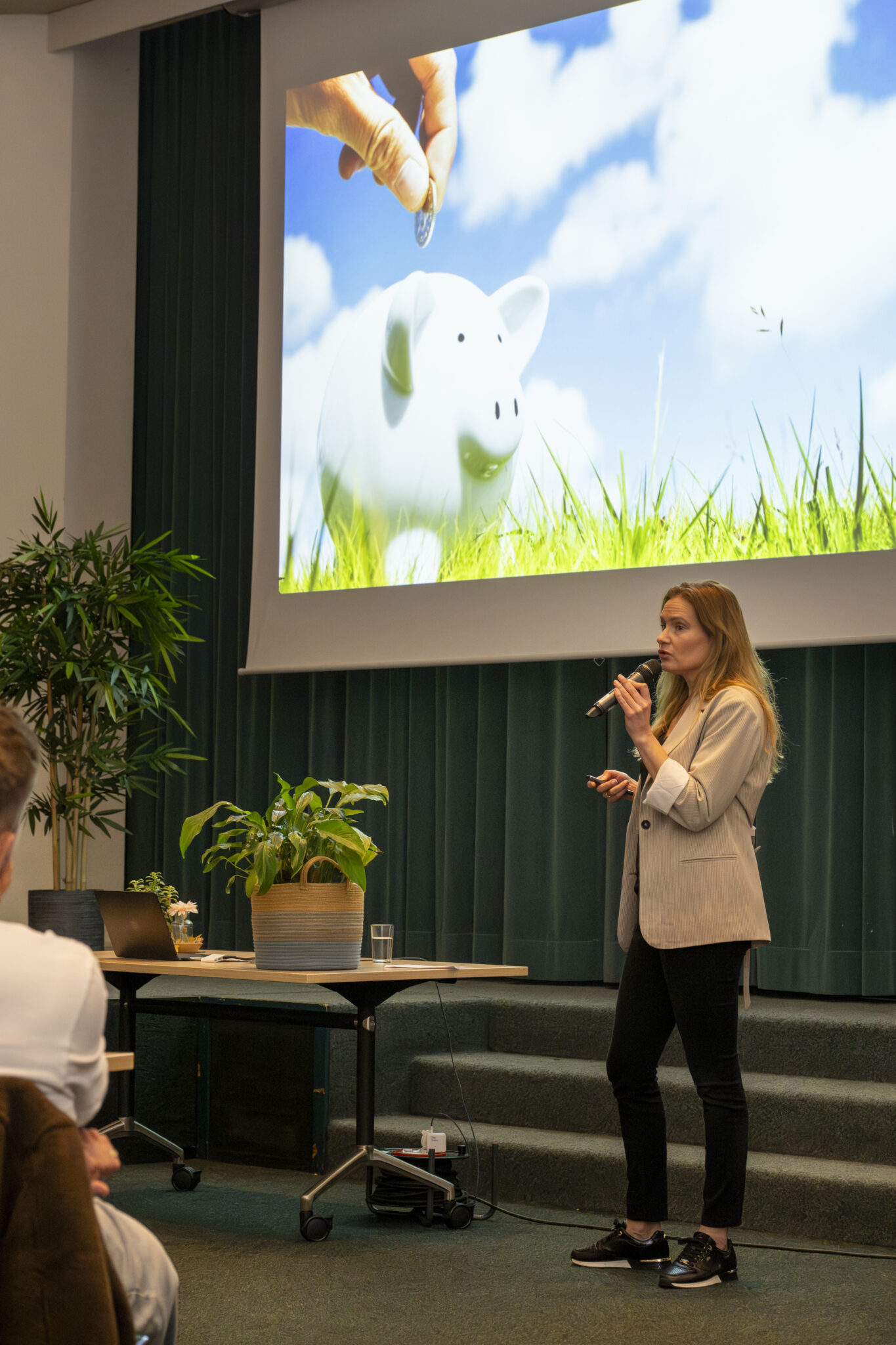 Vrouw presenteert met dia van spaarvarken en munt, voor publiek, naast bureau met planten.