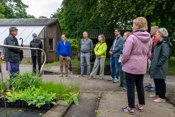 Groep mensen luistert naar een gids op een tuinbouwlocatie naast een kas.