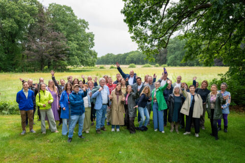 Groep mensen poseert vrolijk in een groen park met bomen en weiland op de achtergrond.