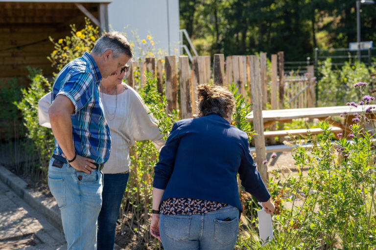 Drie mensen inspecteren planten in een tuin met houten hek en picknicktafel op de achtergrond.