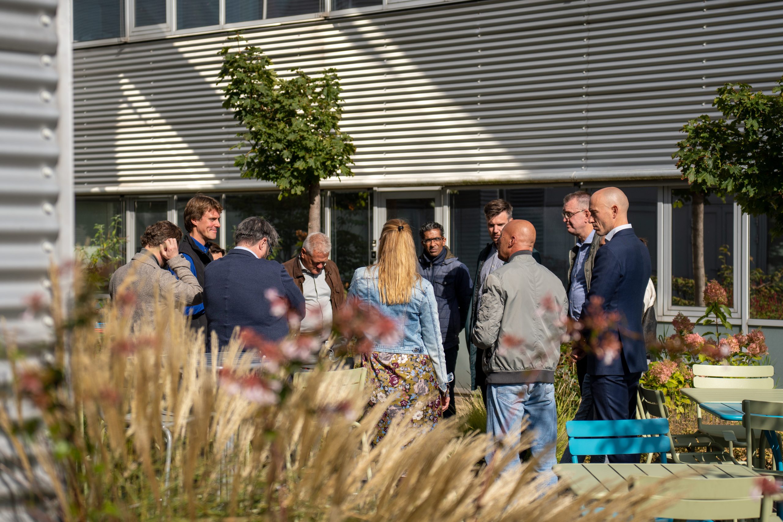 Groep mensen staat in een cirkel buiten bij een gebouw met bomen en bloemen op voorgrond.