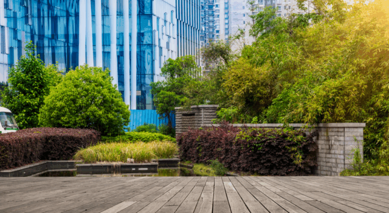 Stedelijke tuin met houten dek, groene struiken en moderne glazen gebouwen op de achtergrond.
