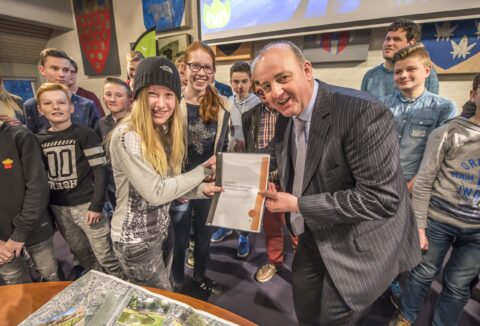 Groep tieners overhandigt rapport aan glimlachende man in pak; kamer vol wanddecoraties.