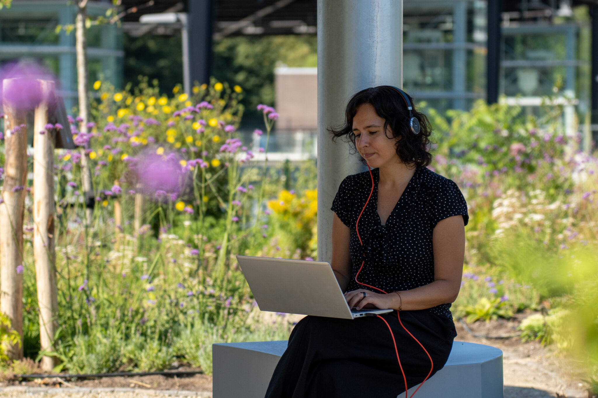 Vrouw met koptelefoon werkt op laptop in een tuin met paarse bloemen.