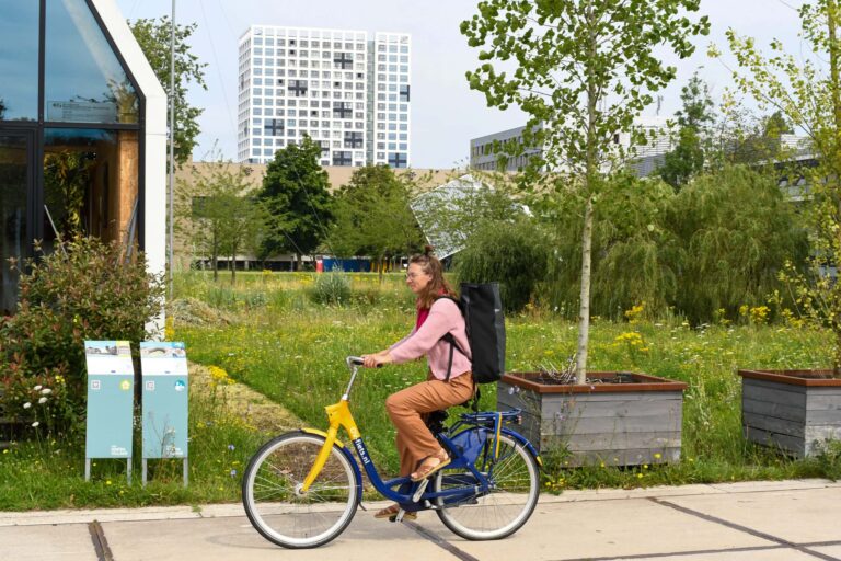 Vrouw fietst met rugtas langs groen park met gebouwen op de achtergrond.