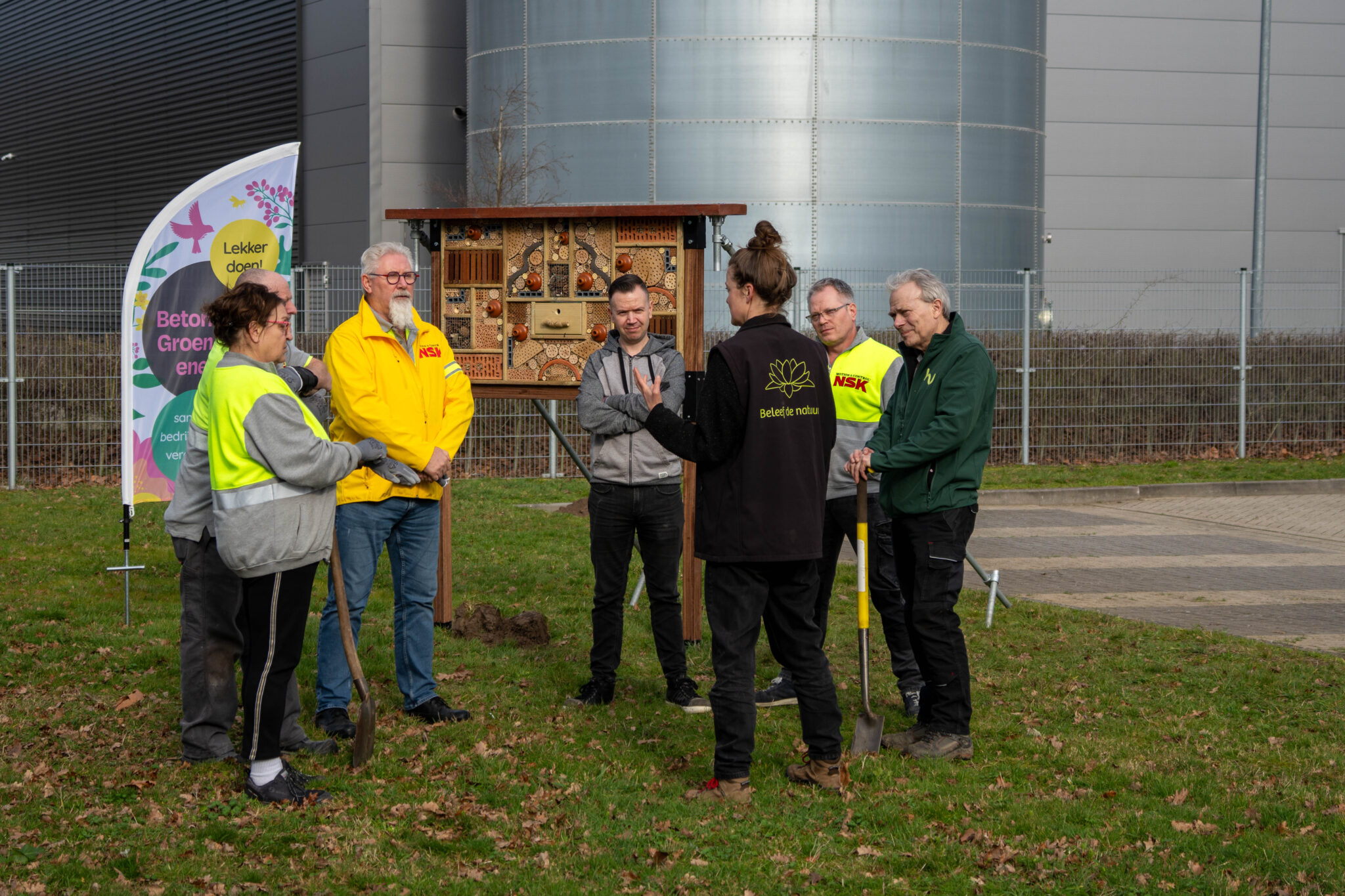 Groep mensen met spades bij een insectenhotel en vlag op grasveld.