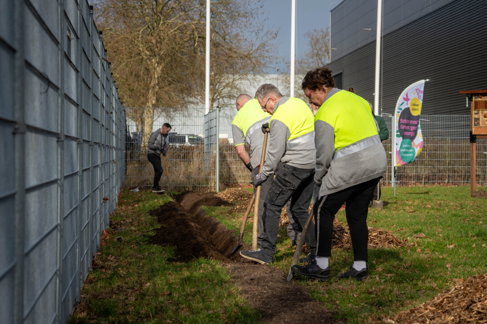 Mensen graven een geul langs een hek, dragen reflecterende veiligheidshesjes.