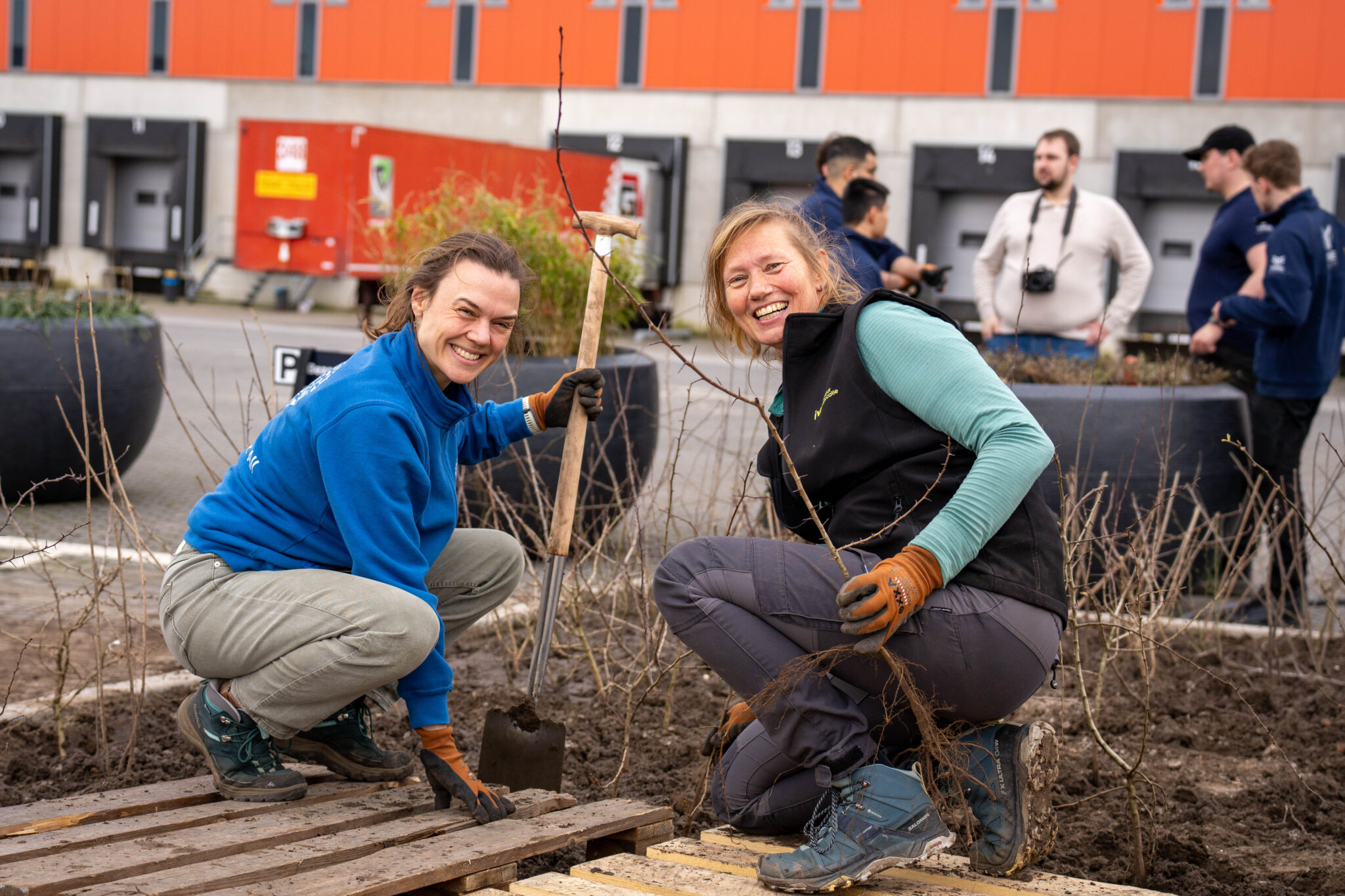 Twee lachende vrouwen planten struiken op een industrieel terrein met oranje gebouw op de achtergrond.