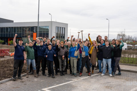 Groep mensen juicht, met vuisten in de lucht, voor een modern gebouw.