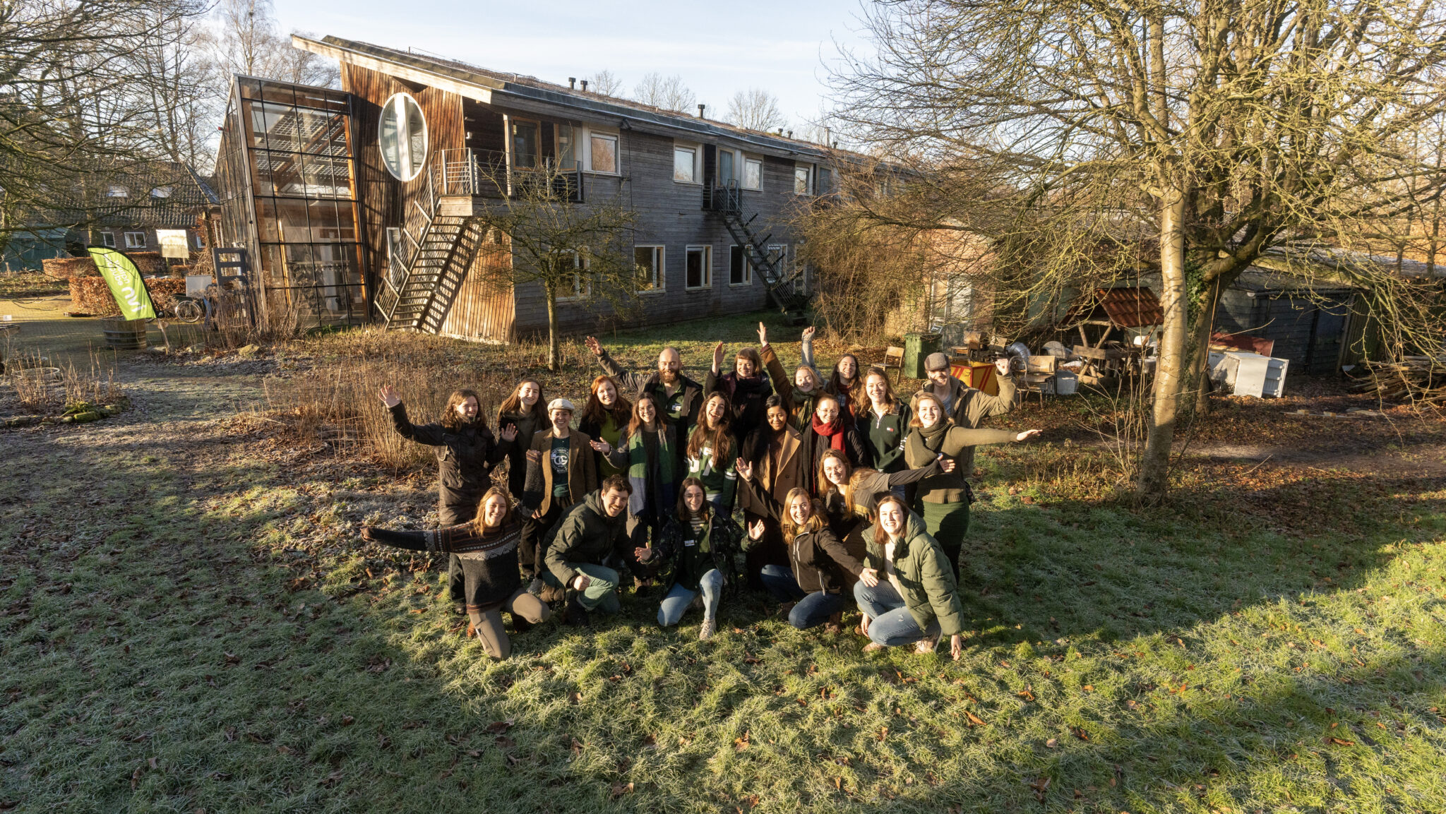 Groep mensen poseert voor houten gebouw met grote ronde raam, omringd door winterse tuin.