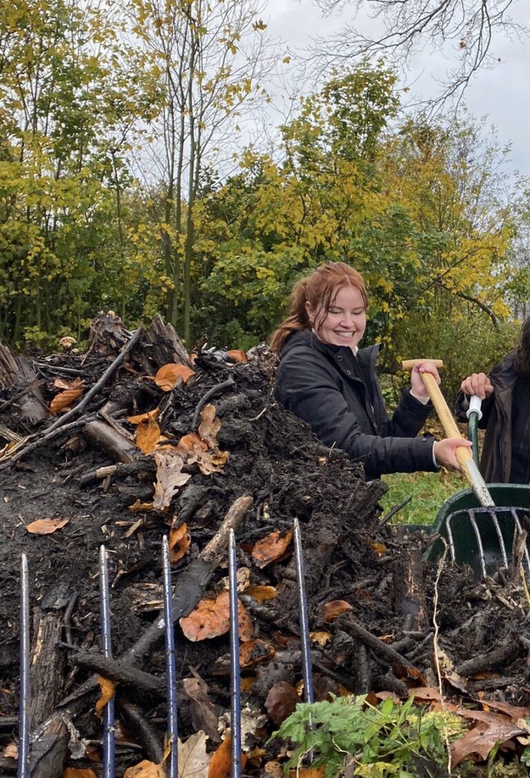 Persoon werkt lachend in de tuin met een hooivork, bladeren en takken op de achtergrond.