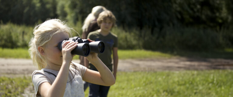 Een kind kijkt door een verrekijker in een groen, zonnig landschap.