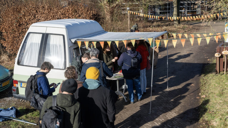 Rij mensen bij een kraampje naast een witte bus met gekleurde vlaggen in een buitenomgeving.