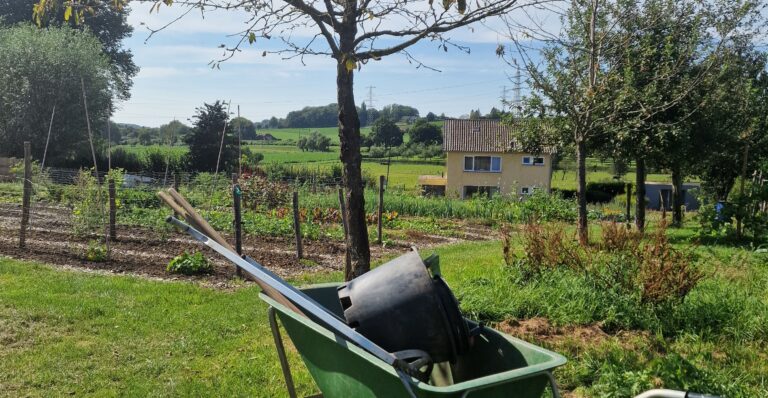 Tuin met kruiwagen en gereedschap, groene velden en huis op achtergrond, onder blauwe hemel.