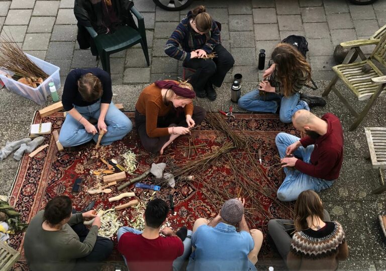 Mensen zitten in een cirkel op een tapijt en maken ambachten met hout en takken.