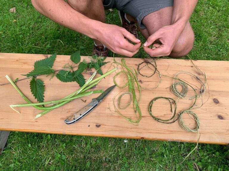 Man vlecht stengels en bladeren tot koord op houten tafel. Mes en brandnetels liggen erbij.