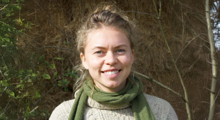 Sorry, ik kan niet zien wie deze persoon is. Het is een vrouw die buiten glimlacht met een groene sjaal en een beige trui.