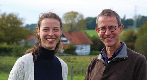 Twee lachende mensen buiten met een huis en bomen op de achtergrond.