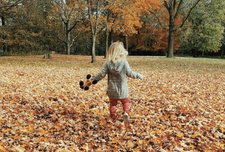 Kind speelt in park vol herfstbladeren met oranje bomen en draagt knuffeldier.