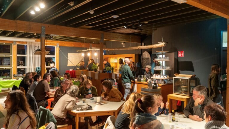 Een drukke cafetaria met mensen die aan tafels zitten en een bar met personeel op de achtergrond.
