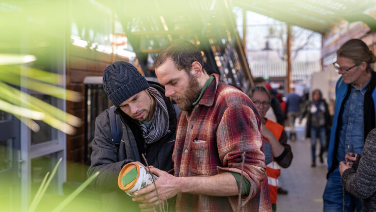 Twee mannen inspecteren samen een object in een drukke binnentuin.