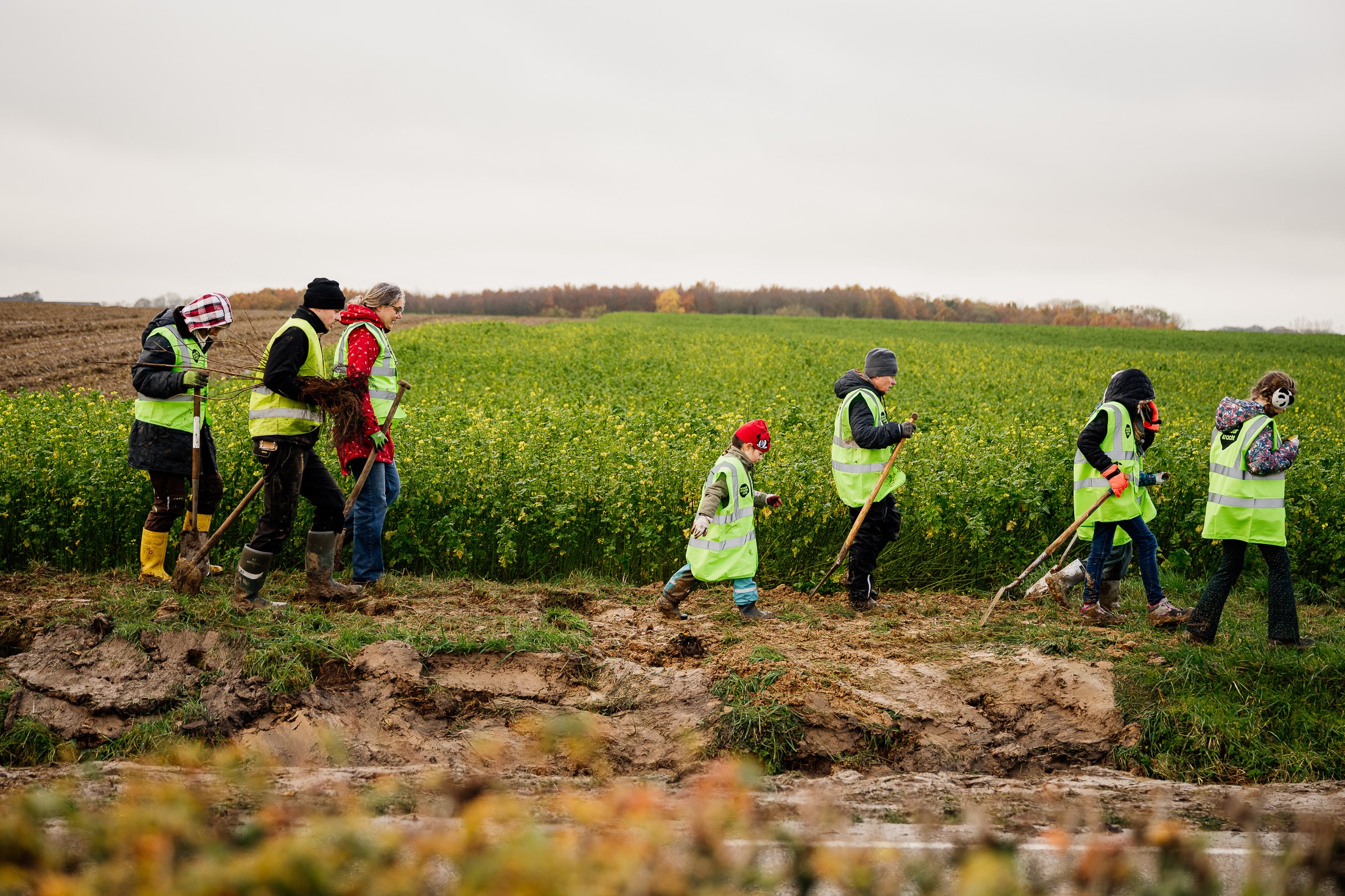 Groep mensen in reflecterende jassen loopt met gereedschap langs een groen veld en modderige grond.