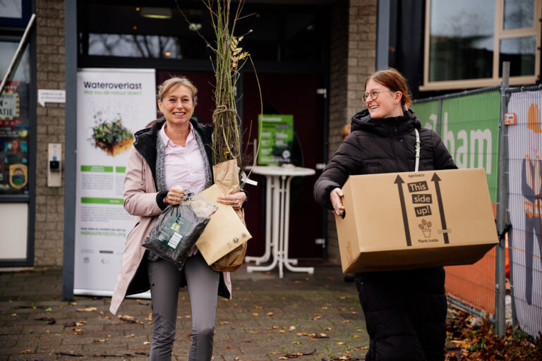 Twee vrouwen dragen planten en een doos voor een gebouw met een banner over waterbeheer.