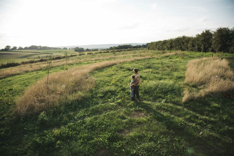 Twee personen omhelzen elkaar op een groen veld met bomen en een heldere lucht op de achtergrond.
