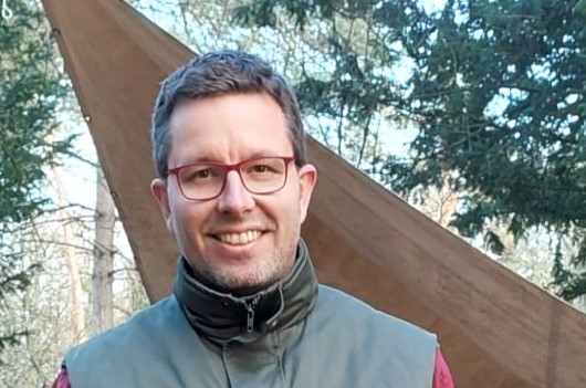 Een glimlachende man met een bril in een groene jas in een bosrijke omgeving.