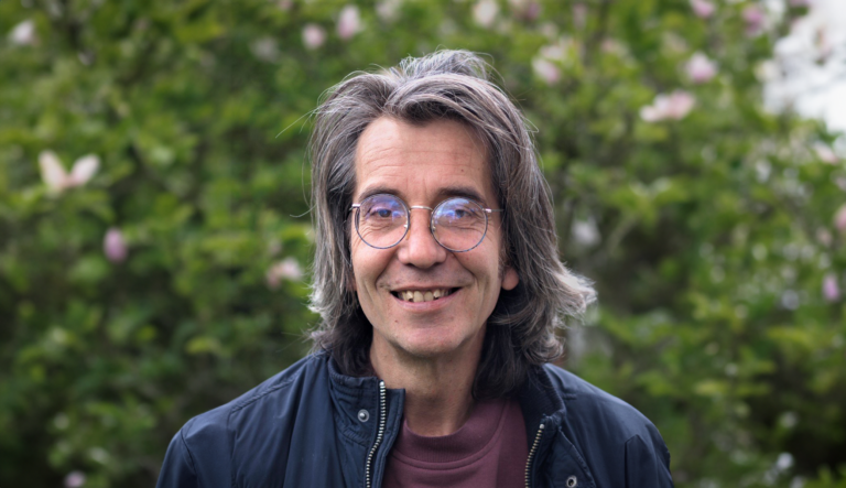 Man met bril en lang haar glimlacht, groene achtergrond met wazige bloemen.