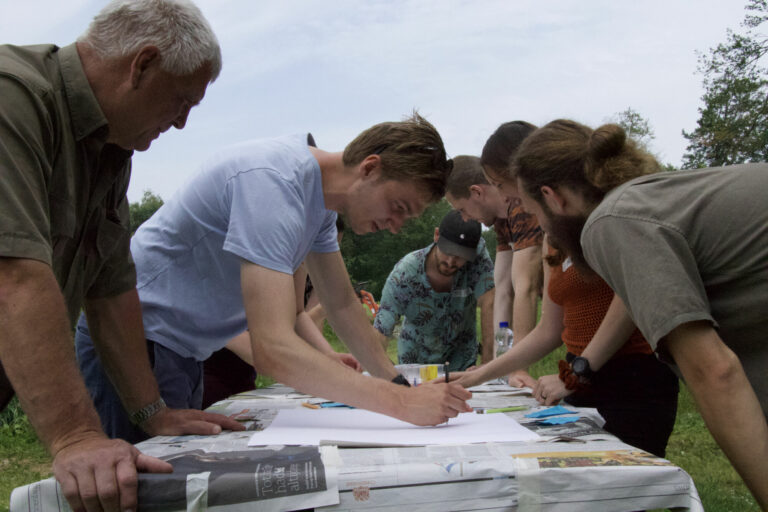 Mensen buigen zich over een tafel buiten en tekenen samen op papier.