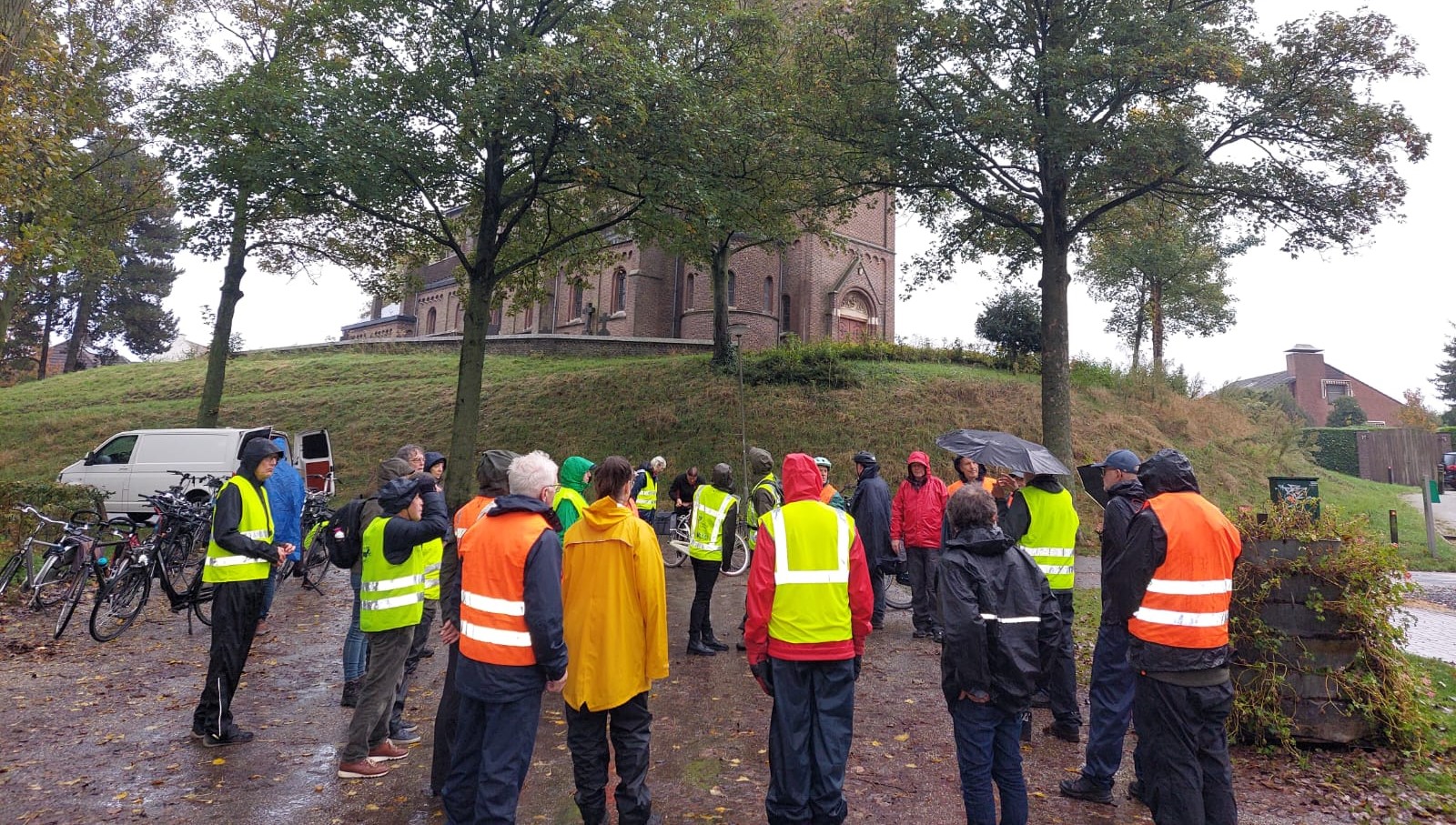 Groep mensen in regenjassen en hesjes bij fietsen, voor een kerk, in regenachtig weer.