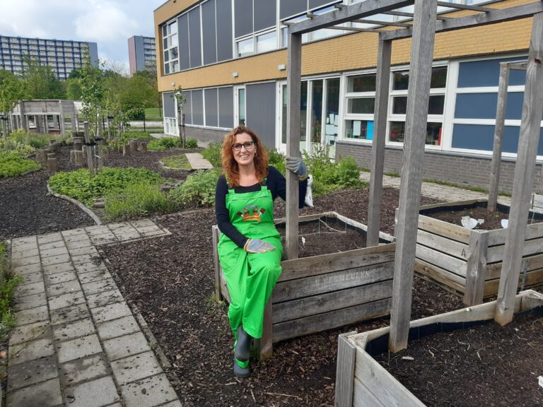 Vrouw in groene overall zit in een schooltuin met houten plantenbakken.