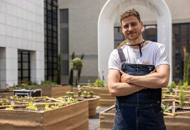 Man in tuinbroek bij verhoogde groentebakken in een binnenplaats met modern gebouwen.