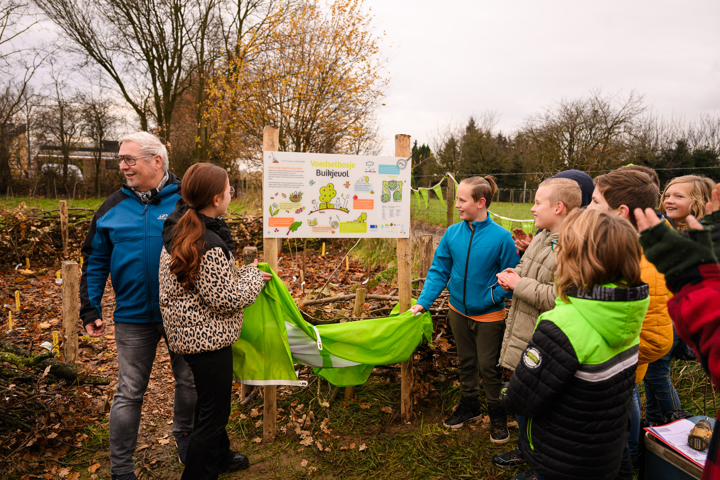 Groep mensen onthult een bord met "Voedselbosje Buikjevol" in een groene buitenomgeving.