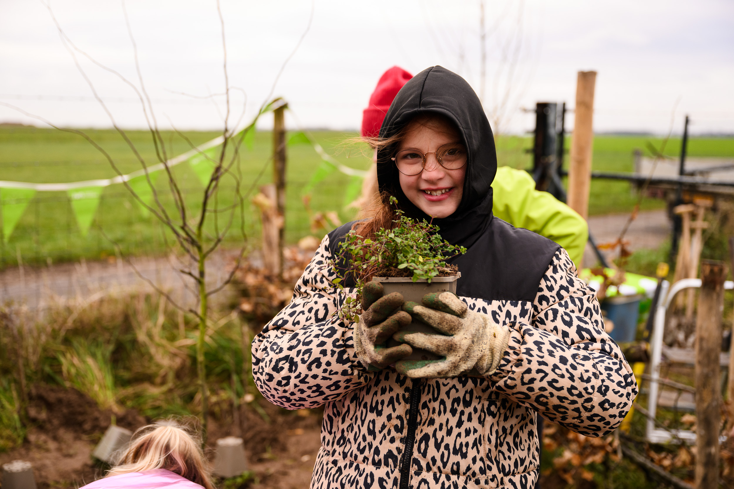 Meisje met bril in luipaardjas houdt plant vast in een tuin, achtergrond met groene velden.