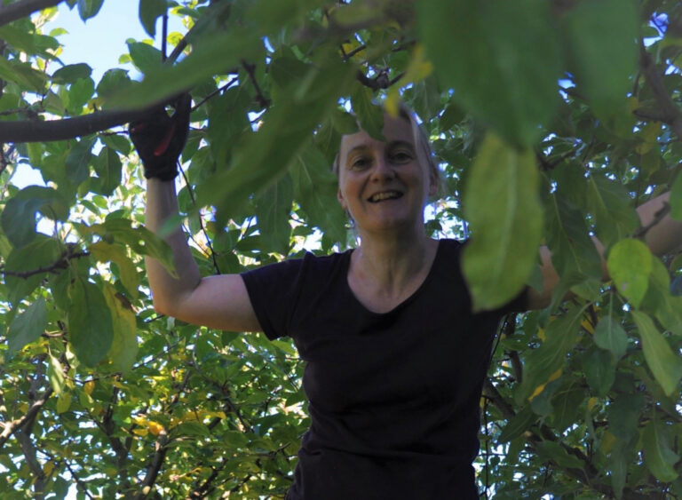 Persoon glimlachend tussen groene bladeren, gekleed in een zwarte shirt, werkomgeving buiten.