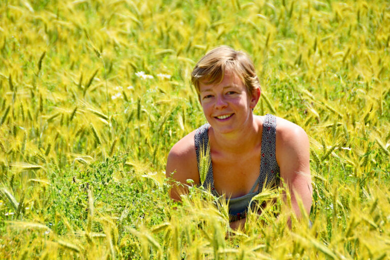 Vrouw glimlacht in een weelderig veld met geelgroen graan en enkele witte bloemen.