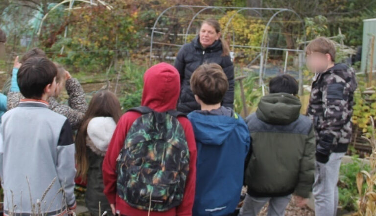 Groep kinderen luistert naar een vrouw in een tuin met tuinkassen.
