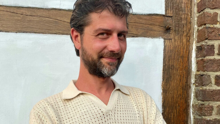 Man met baard glimlacht voor witte muur met houten balk en bakstenen.