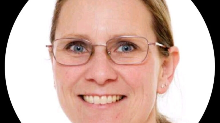 Portret van een glimlachende vrouw met bril en lichte achtergrond.