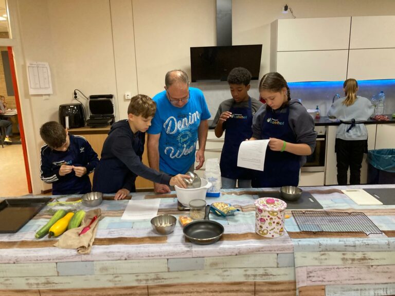 Groep kinderen en een volwassene koken samen in een keuken naast ingrediënten en pannen.