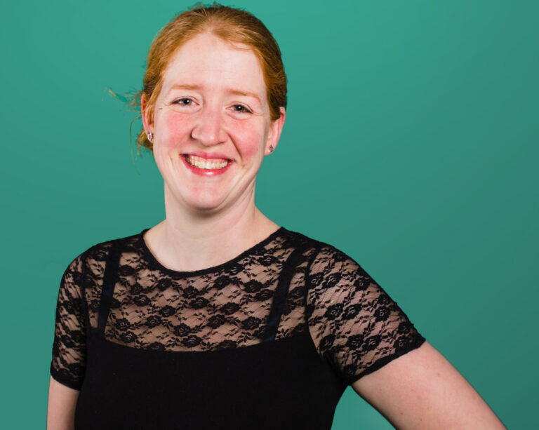 Vrouw met rood haar en zwarte kanten top lacht voor een groene achtergrond.