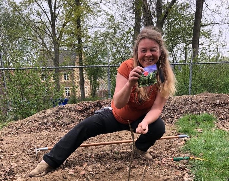 Vrouw in tuin, glimlachend, toont zaadpakket, omgeven door bomen en hekken.