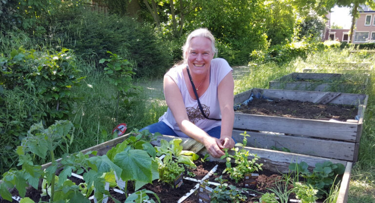 Vrouw in moestuin, zittend en glimlachend, omringd door groenten in verhoogde bakken.
