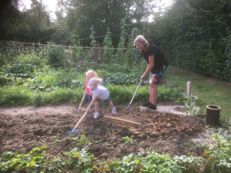 Volwassen vrouw en twee kinderen harken samen in een groene moestuin.