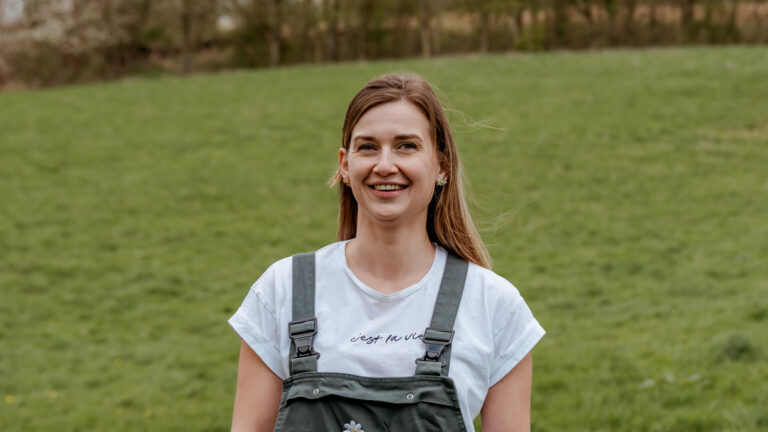 Vrouw glimlacht in een groene weide, draagt tuinbroek en een T-shirt met tekst.