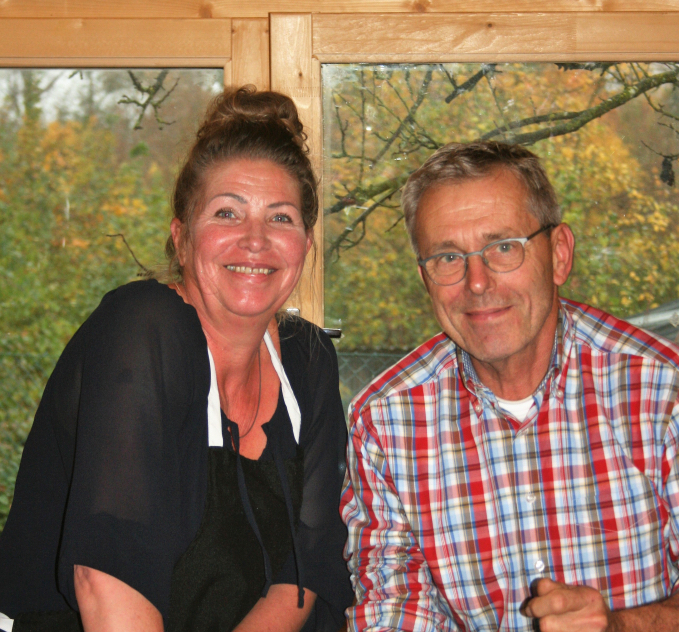 Man en vrouw zitten samen voor een raam met herfstbomen op de achtergrond. Beiden glimlachen.